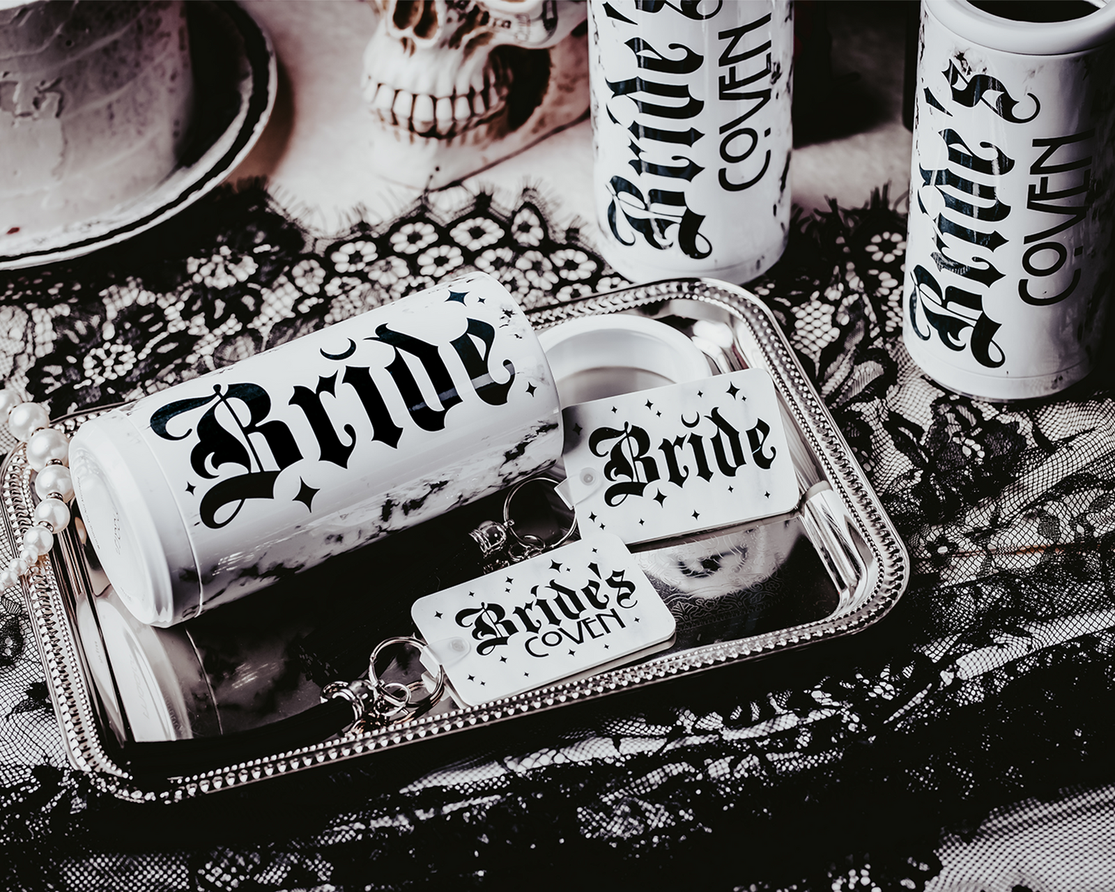 Set of three white, marble-patterned skinny can coolers with gothic black lettering. One cooler reads 'Bride,' and the other two read 'Bride's Coven,' all set against a dark, moody background with lace, disco balls, and red roses, creating a gothic and alternative bridal party theme.