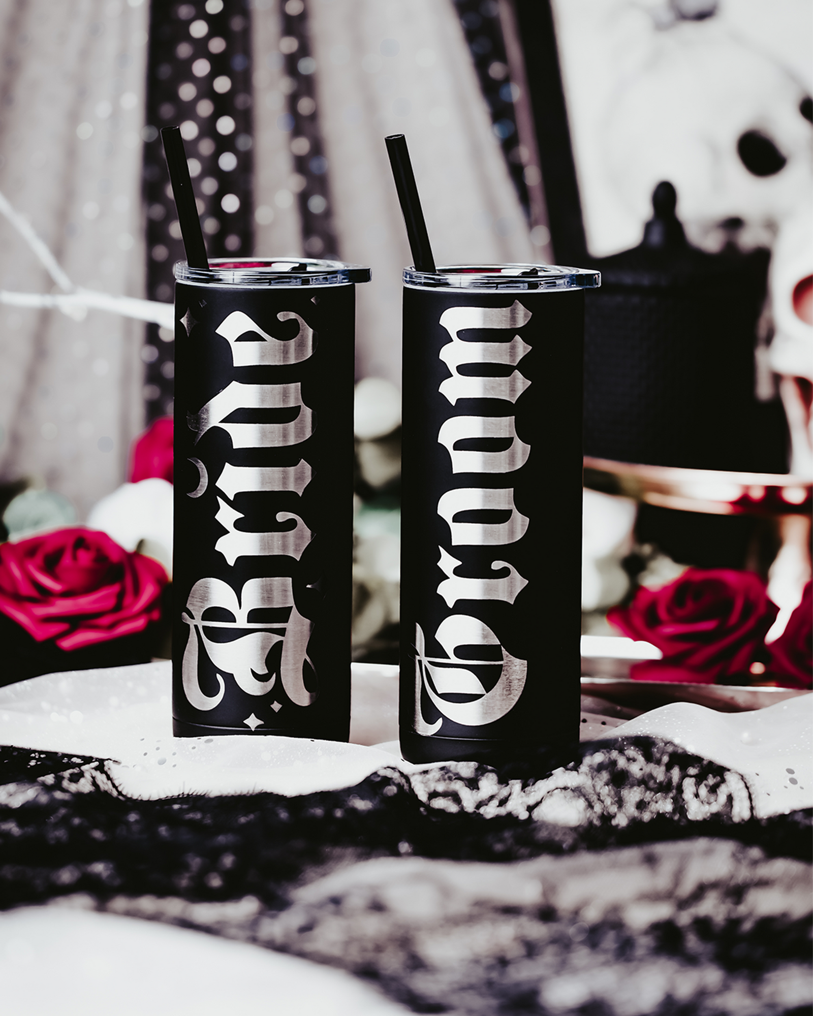 Set of two black stainless steel tumblers, one engraved with 'Bride' and the other with 'Groom' in bold, gothic silver lettering. Each tumbler features a black straw and is set against a dramatic, gothic backdrop with red roses, lace, and soft, romantic lighting, perfect for an alternative or darkly romantic wedding theme.