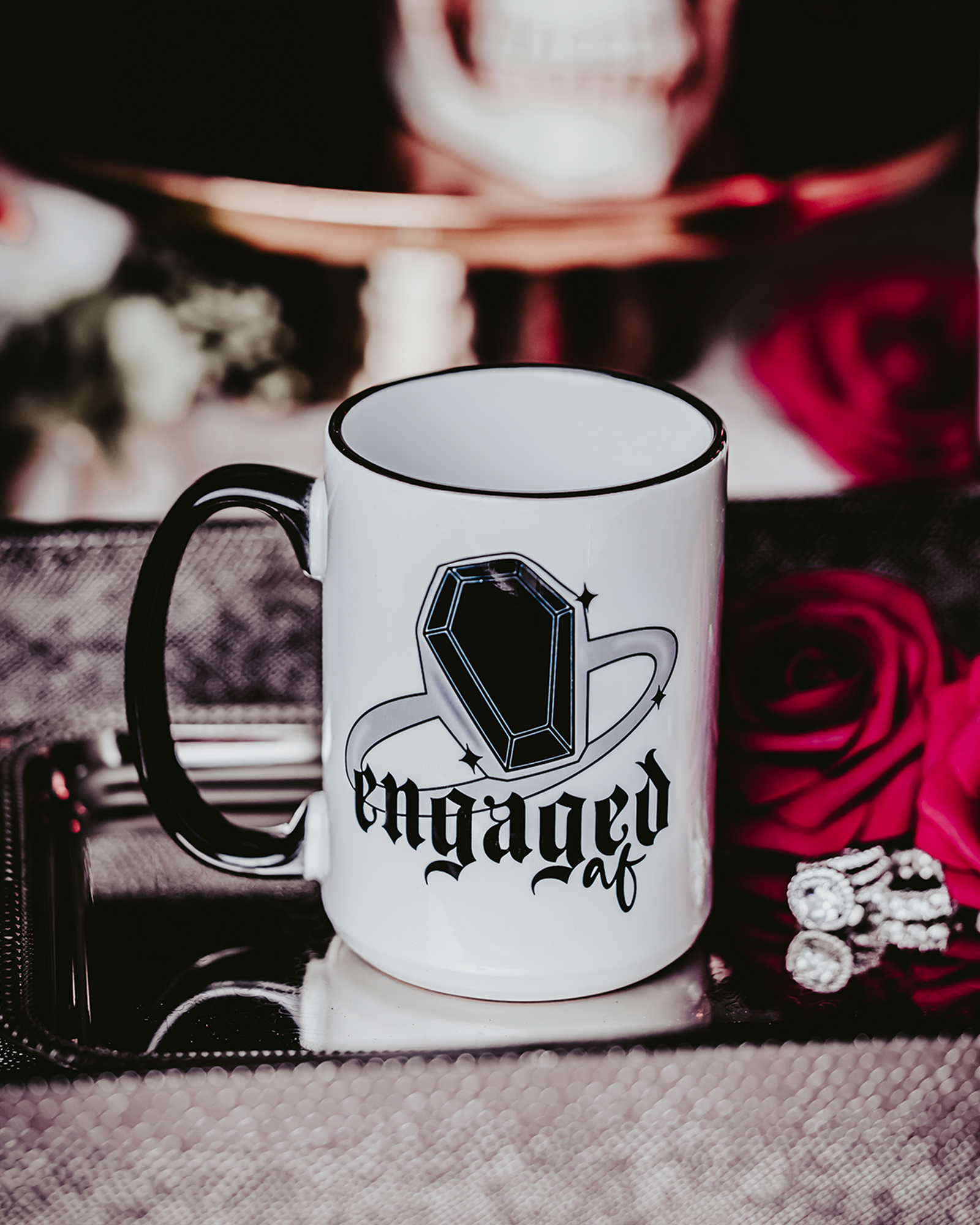 A white ceramic mug with a black handle and rim, featuring a bold black gem ring design and the phrase "Engaged AF" in gothic-style font. The mug is displayed against a dark, romantic setting with red roses in the background, capturing an edgy and gothic bridal theme.