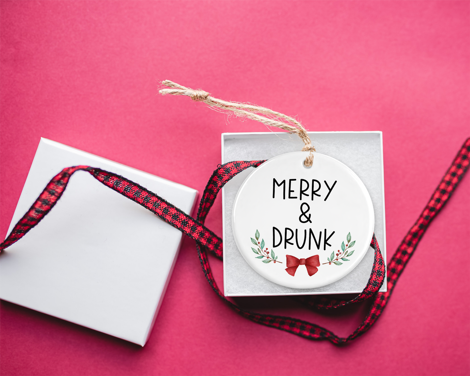 White ceramic ornament with the phrase "Merry & Drunk" adorned with holiday greenery and a red bow, hanging by twine in a festive gift box.