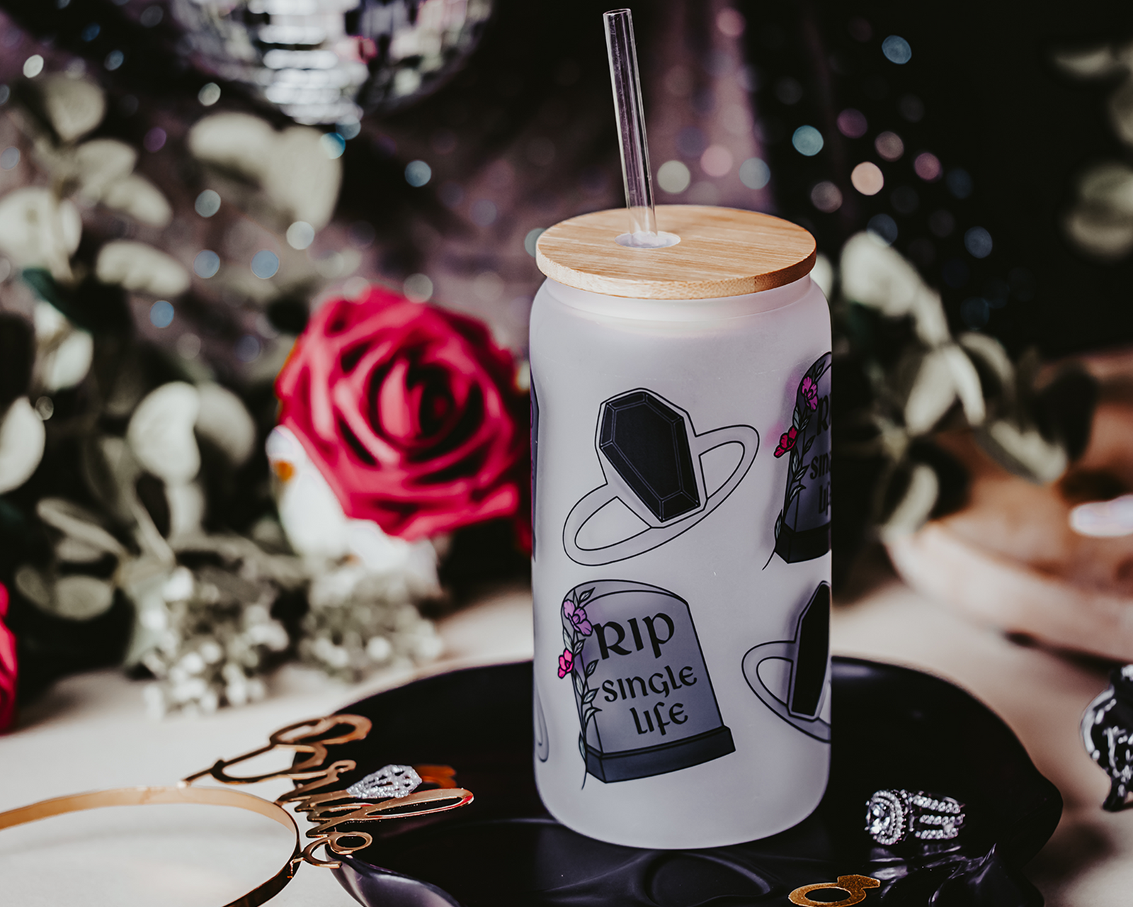 Frosted glass can with a bamboo lid and straw, featuring gothic-themed illustrations of a coffin and tombstone with the phrase 'RIP Single Life.' The can is surrounded by red and white roses, a gold 'Bride' headband, and decorative rings, creating a dark, romantic bridal atmosphere.