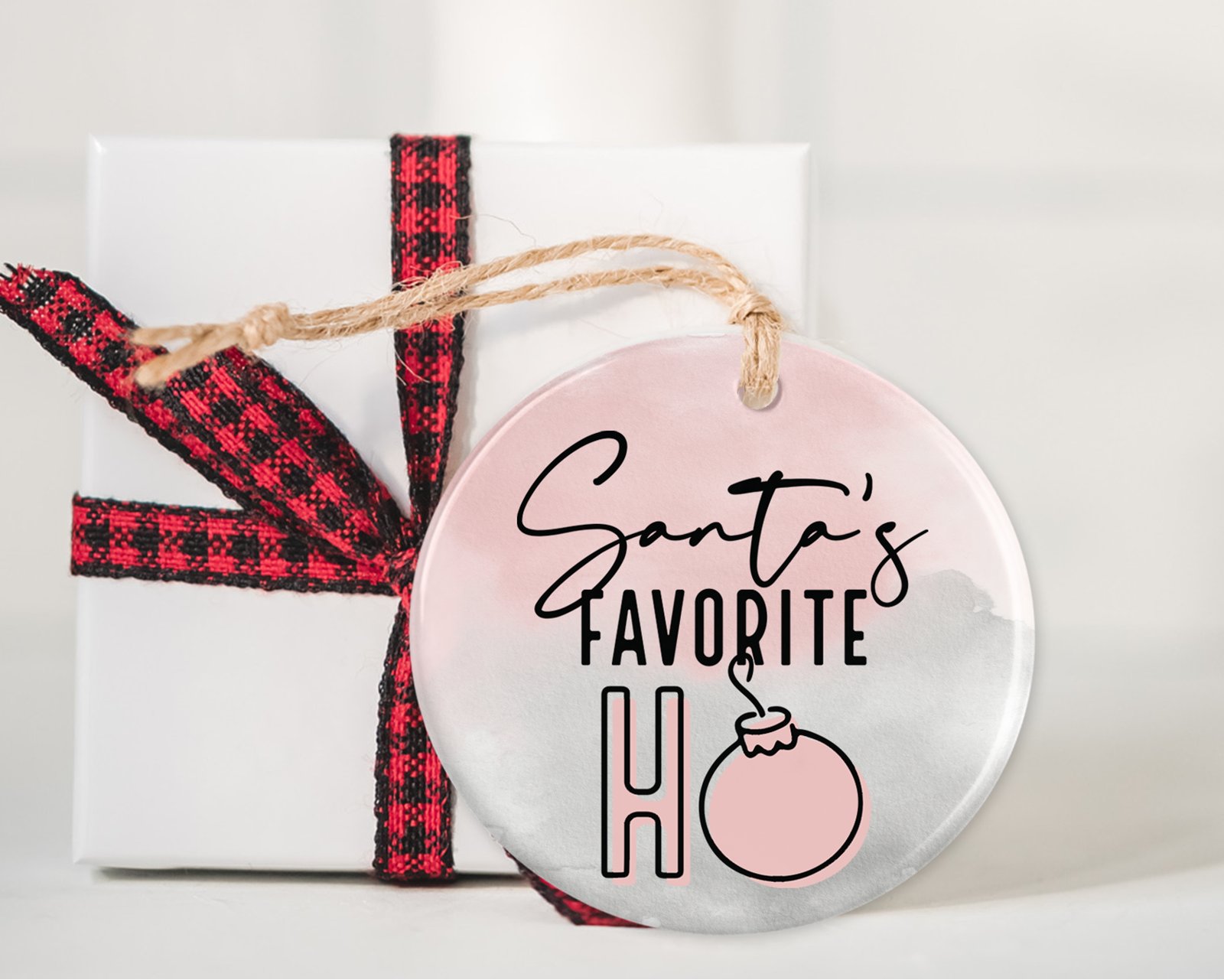 Pink and white ceramic ornament with "Santa's Favorite Ho" written in playful black and pink lettering, hanging on a twine string, placed next to a white gift box with a red and black plaid ribbon.