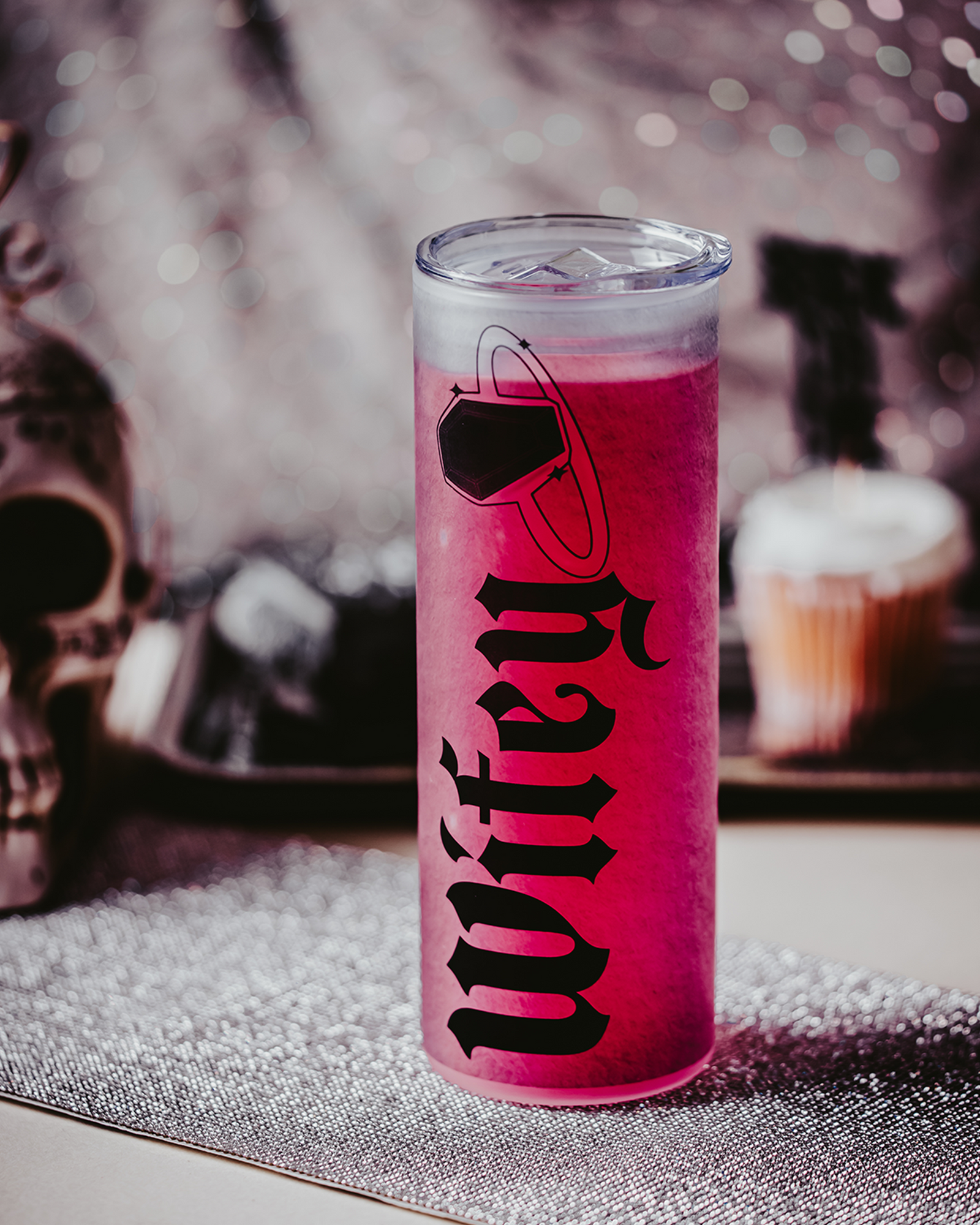 Frosted glass tumbler with gothic black lettering spelling 'Wifey,' accompanied by a coffin ring illustration. The tumbler is lying on its side amidst red roses, cupcakes, and decorative elements, creating a dark, romantic bridal vibe.