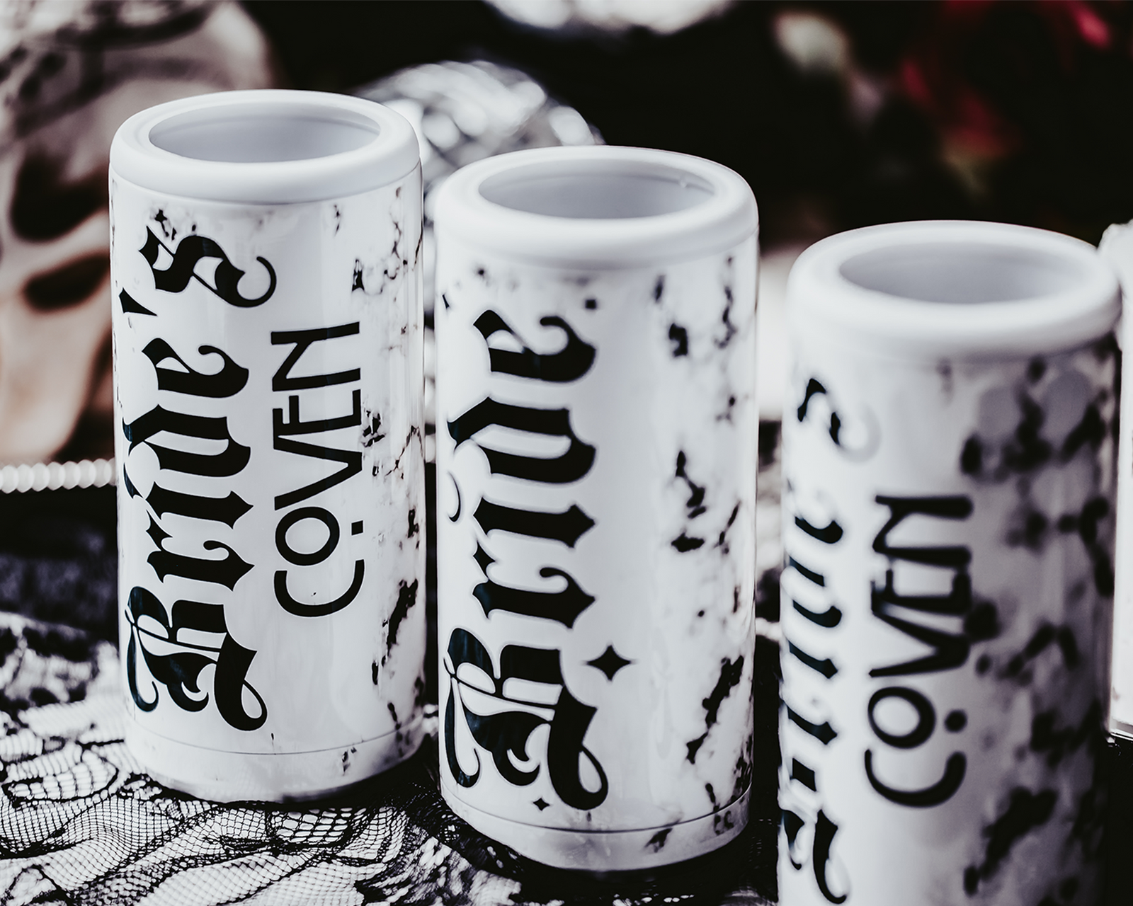 Set of three white, marble-patterned skinny can coolers with gothic black lettering. One cooler reads 'Bride,' and the other two read 'Bride's Coven,' all set against a dark, moody background with lace, disco balls, and red roses, creating a gothic and alternative bridal party theme.