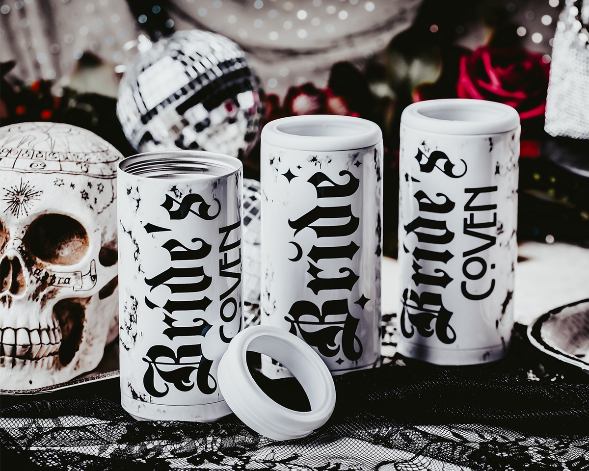 Set of three white, marble-patterned skinny can coolers with gothic black lettering. One cooler reads &#39;Bride,&#39; and the other two read &#39;Bride&#39;s Coven,&#39; all set against a dark, moody background with lace, disco balls, and red roses, creating a gothic and alternative bridal party theme.