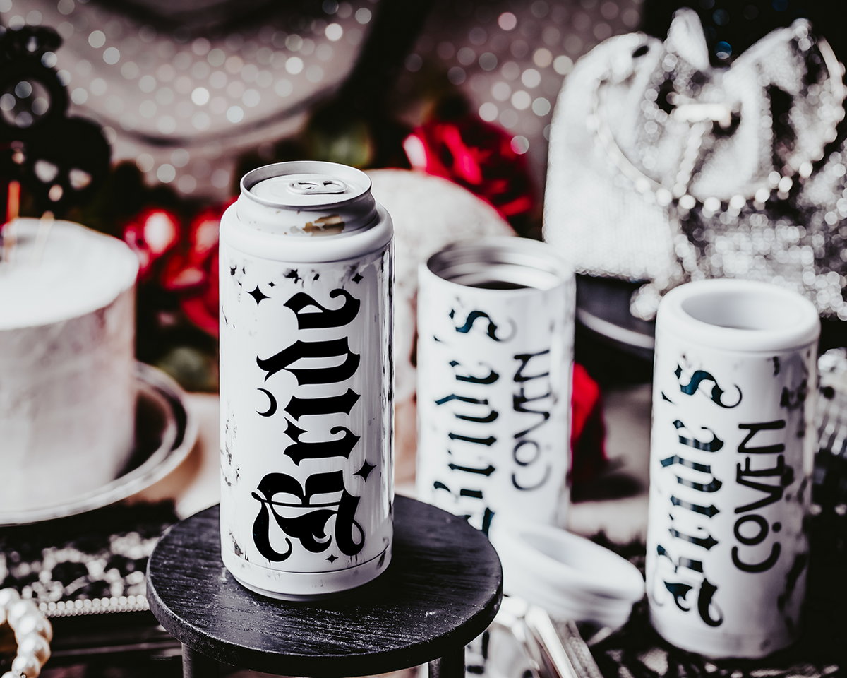 Set of three white, marble-patterned skinny can coolers with gothic black lettering. One cooler reads &#39;Bride,&#39; and the other two read &#39;Bride&#39;s Coven,&#39; all set against a dark, moody background with lace, disco balls, and red roses, creating a gothic and alternative bridal party theme.