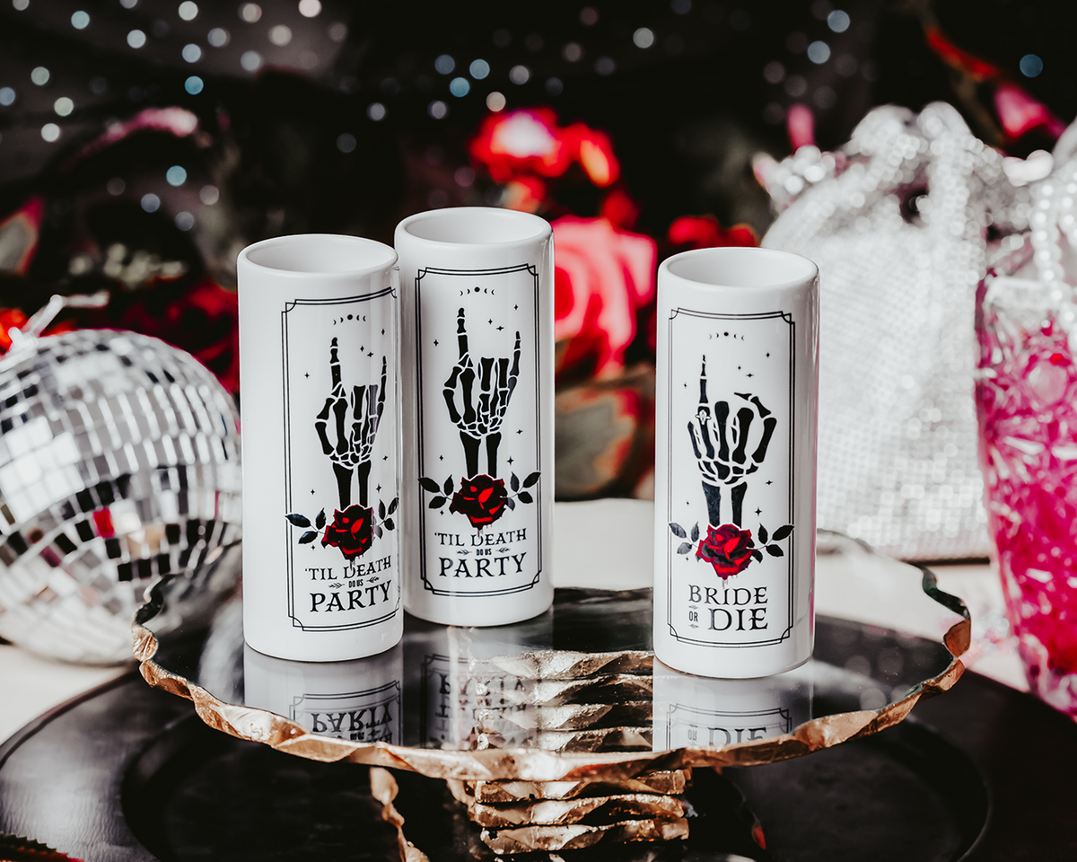 Gothic-style ceramic shot glasses featuring skeleton hand designs with red roses and phrases like &quot;Bride or Die&quot; and &quot;&#39;Til Death Do Us Party,&quot; perfect for an alternative bridal party.