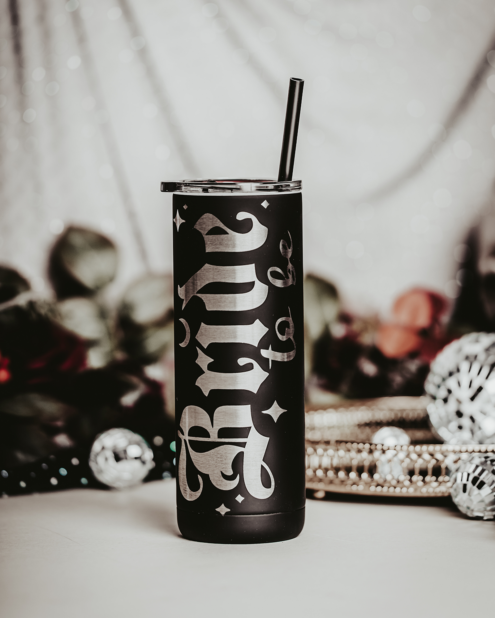 Black stainless steel tumbler with the phrase 'Bride to be' in bold, gothic silver lettering. The tumbler features a black straw and is set against a backdrop of soft, bokeh lights with decorative elements, including flowers and small disco balls, creating an elegant and festive bridal scene.