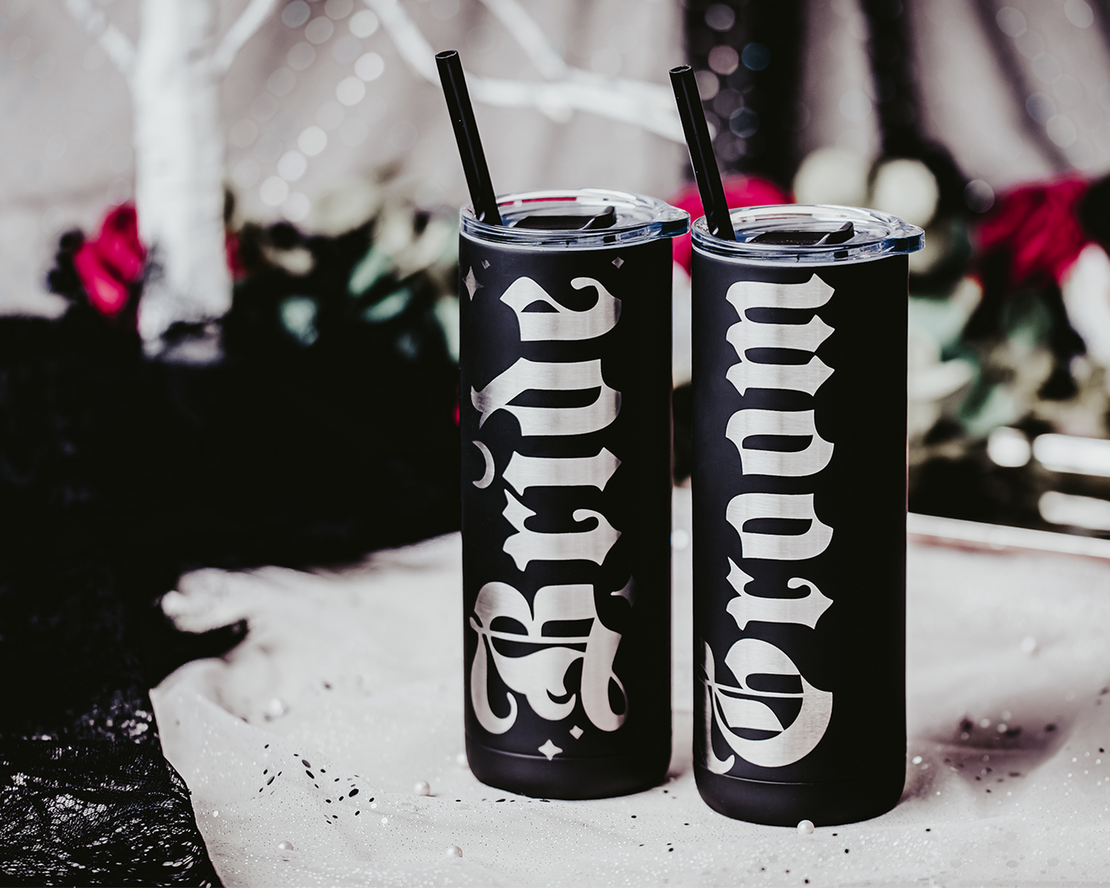 Set of two black stainless steel tumblers, one engraved with 'Bride' and the other with 'Groom' in bold, gothic silver lettering. Each tumbler features a black straw and is set against a dramatic, gothic backdrop with red roses, lace, and soft, romantic lighting, perfect for an alternative or darkly romantic wedding theme.
