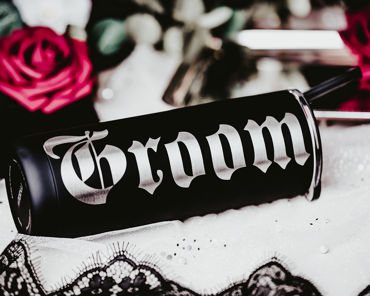 Set of two black stainless steel tumblers, one engraved with &#39;Bride&#39; and the other with &#39;Groom&#39; in bold, gothic silver lettering. Each tumbler features a black straw and is set against a dramatic, gothic backdrop with red roses, lace, and soft, romantic lighting, perfect for an alternative or darkly romantic wedding theme.