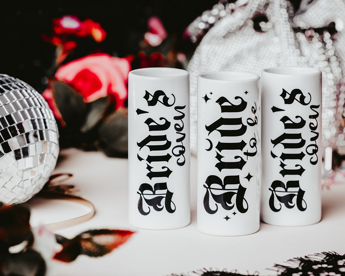 Ceramic shot glasses with gothic-style &#39;Bride to Be&#39; and &#39;Bride&#39;s Coven&#39; typography, perfect for a non-traditional bride and her bridal party, set against a dark, elegant backdrop.