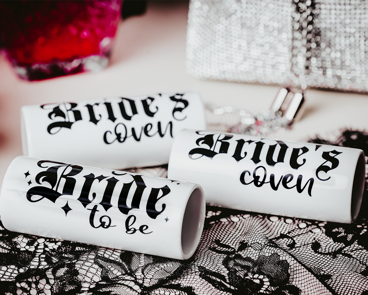 Ceramic shot glasses with gothic-style &#39;Bride to Be&#39; and &#39;Bride&#39;s Coven&#39; typography, perfect for a non-traditional bride and her bridal party, set against a dark, elegant backdrop.
