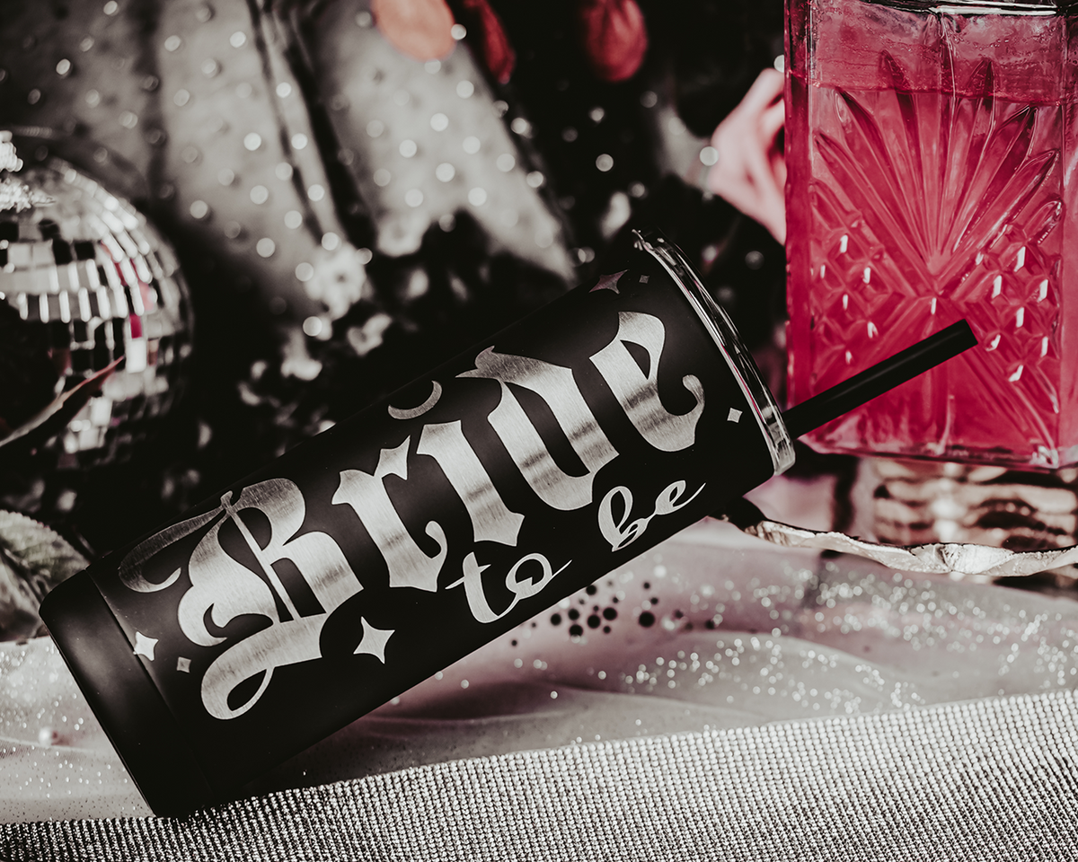 Black stainless steel tumbler with the phrase &#39;Bride to be&#39; in bold, gothic silver lettering. The tumbler features a black straw and is set against a backdrop of soft, bokeh lights with decorative elements, including flowers and small disco balls, creating an elegant and festive bridal scene.
