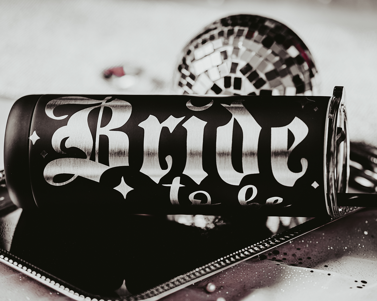 Black stainless steel tumbler with the phrase &#39;Bride to be&#39; in bold, gothic silver lettering. The tumbler features a black straw and is set against a backdrop of soft, bokeh lights with decorative elements, including flowers and small disco balls, creating an elegant and festive bridal scene.