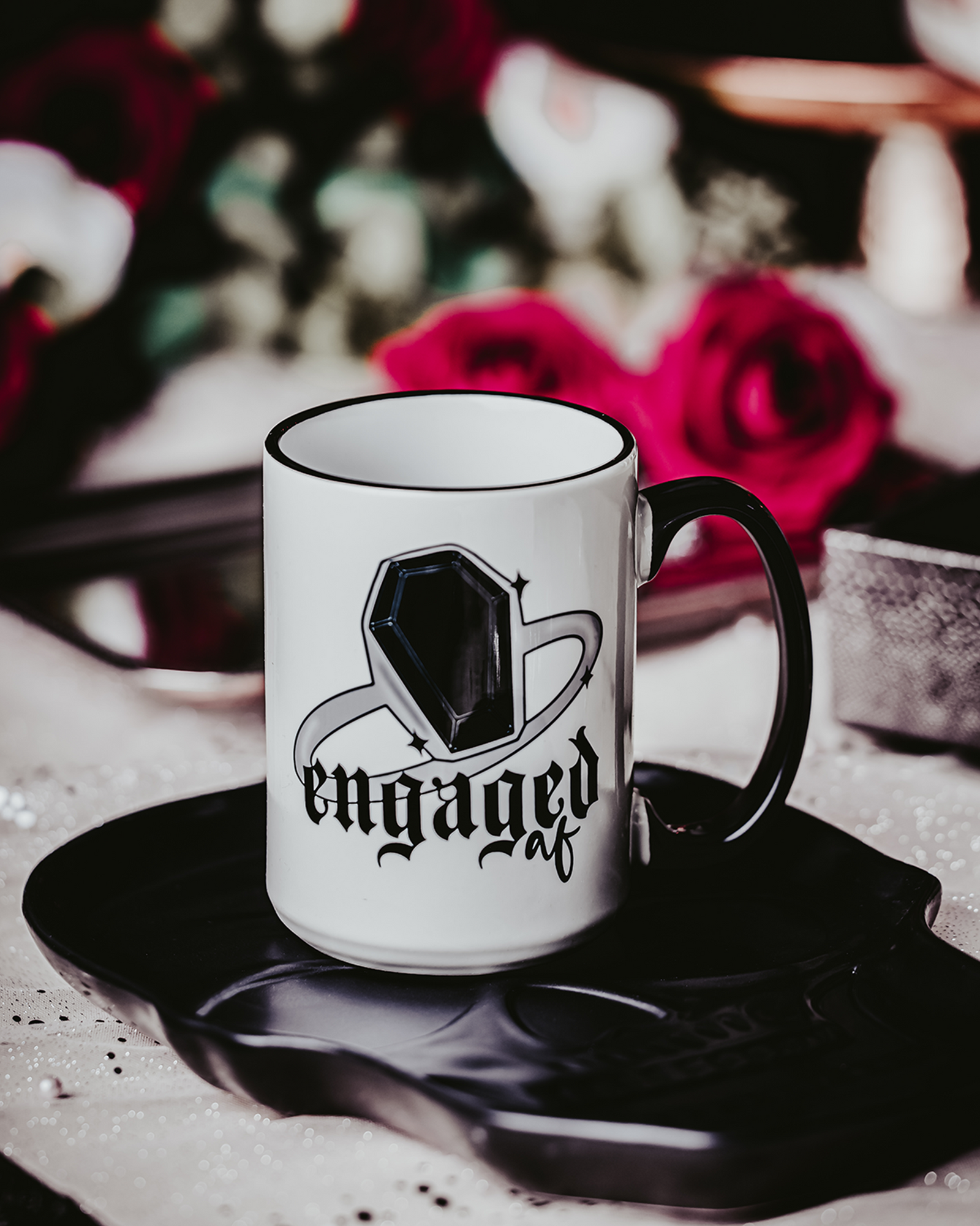 A white ceramic mug with a black handle and rim, featuring a bold black gem ring design and the phrase "Engaged AF" in gothic-style font. The mug is displayed against a dark, romantic setting with red roses in the background, capturing an edgy and gothic bridal theme.