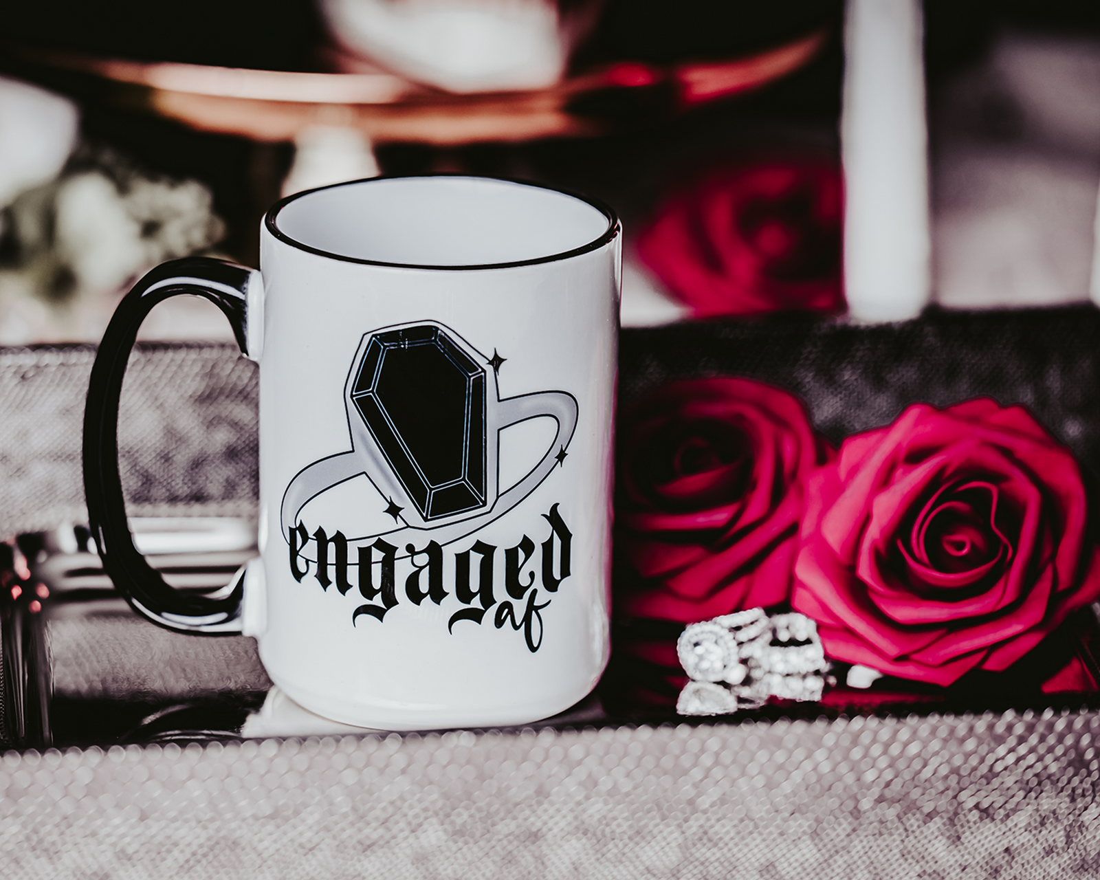  A white ceramic mug with a black handle and rim, featuring a bold black gem ring design and the phrase "Engaged AF" in gothic-style font. The mug is displayed against a dark, romantic setting with red roses in the background, capturing an edgy and gothic bridal theme.