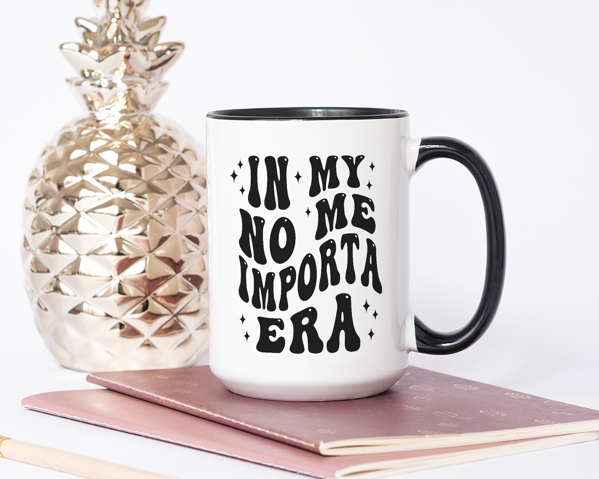 White ceramic mug with a black handle and rim, featuring bold black text that reads &quot;In My No Me Importa Era&quot; in a groovy, retro-style font, surrounded by small decorative sparkles. 