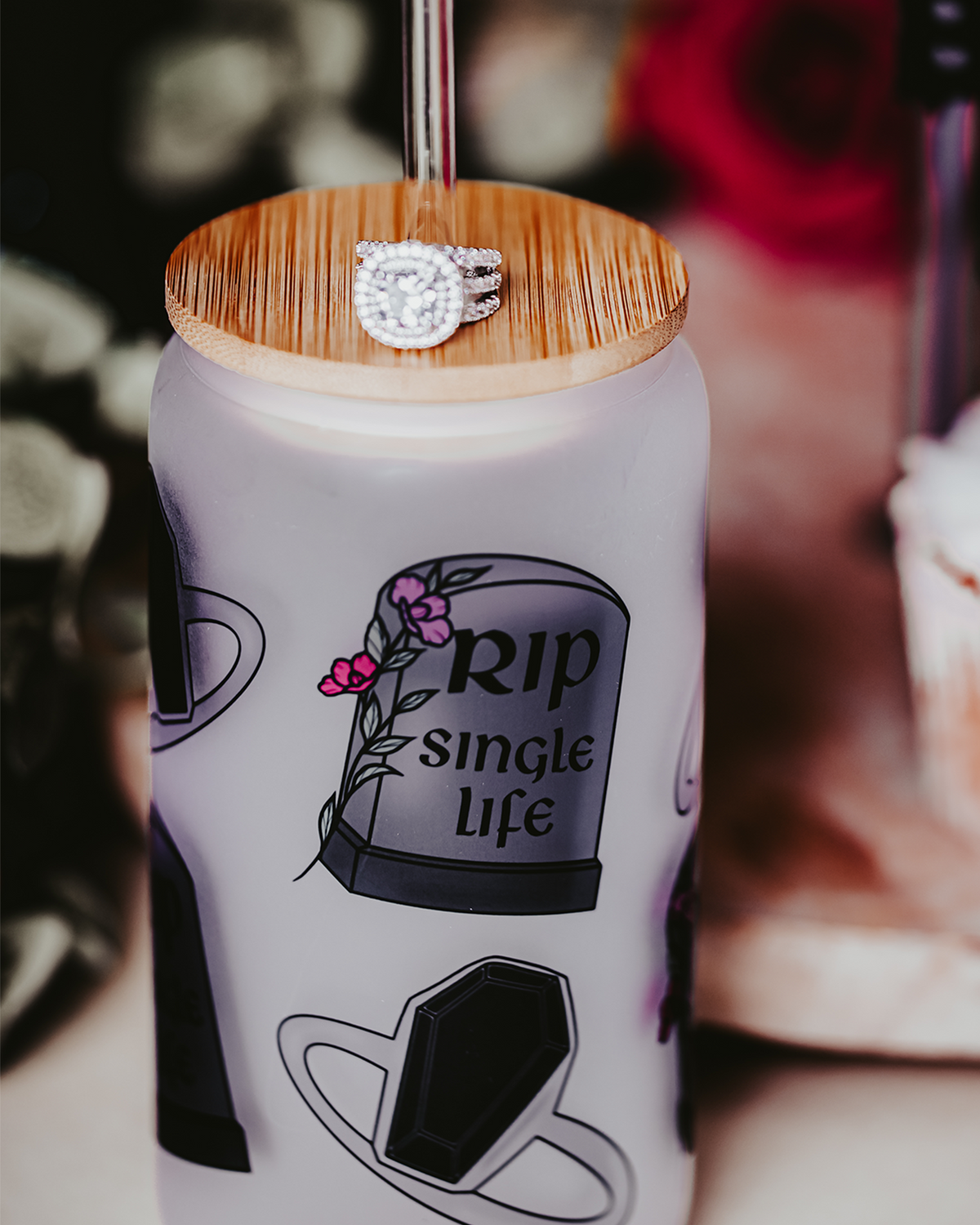 Frosted glass can with a bamboo lid and straw, featuring gothic-themed illustrations of a coffin and tombstone with the phrase &#39;RIP Single Life.&#39; The can is surrounded by red and white roses, a gold &#39;Bride&#39; headband, and decorative rings, creating a dark, romantic bridal atmosphere.