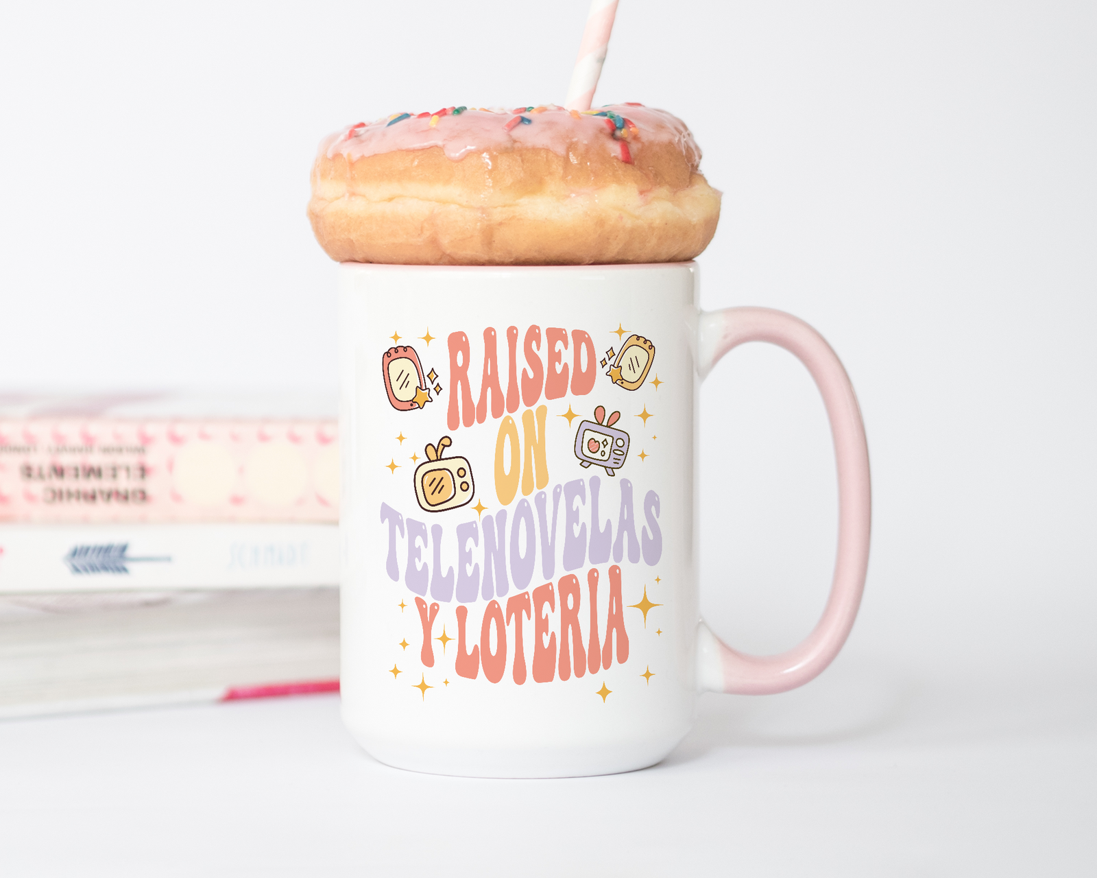 White and pink ceramic mug with retro illustrations and bold text that reads “Raised On Telenovelas Y Lotería” in red, yellow, and purple fonts.