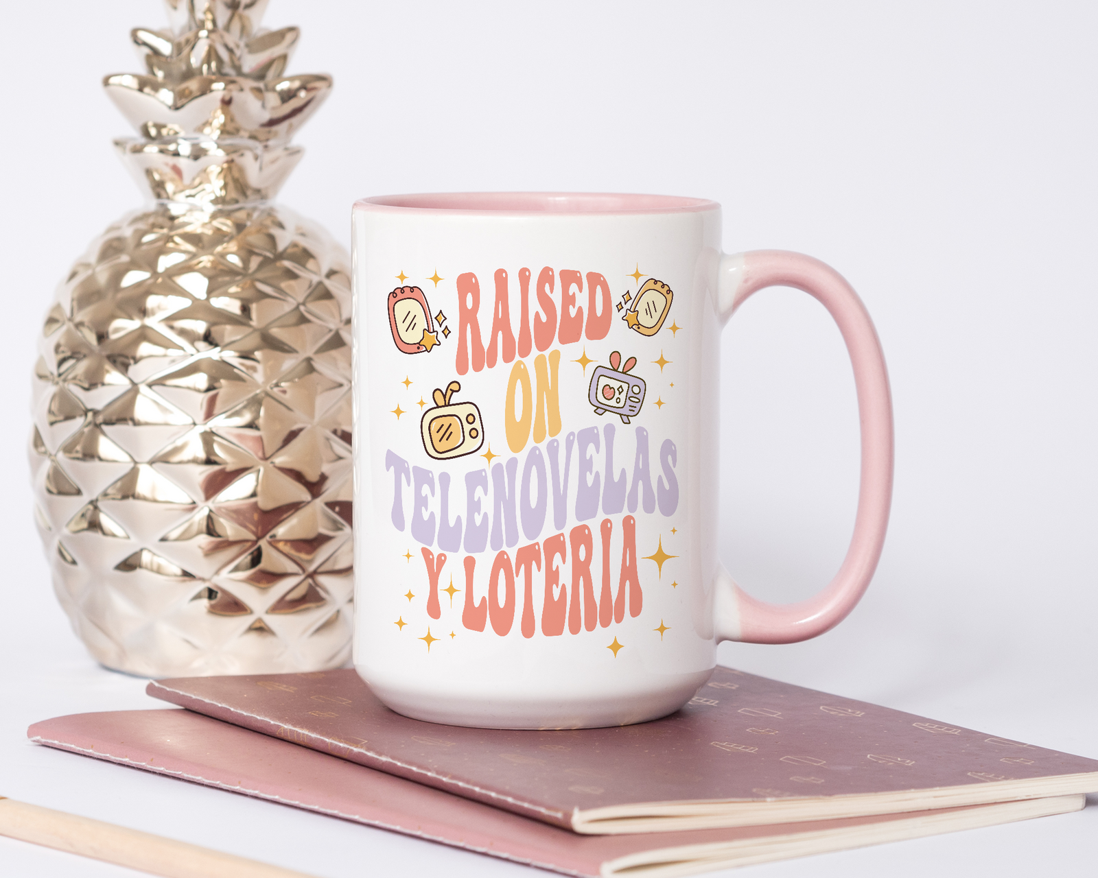 White and pink ceramic mug with retro illustrations and bold text that reads “Raised On Telenovelas Y Lotería” in red, yellow, and purple fonts.