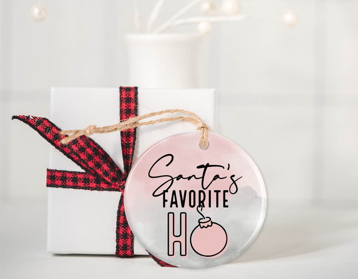 Pink and white ceramic ornament with &quot;Santa&#39;s Favorite Ho&quot; written in playful black and pink lettering, hanging on a twine string, placed next to a white gift box with a red and black plaid ribbon.