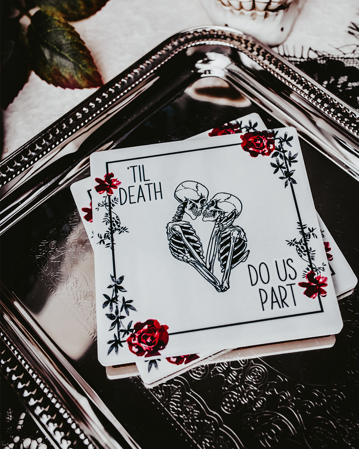 Two square coasters featuring a gothic &#39;Til Death Do Us Part&#39; design with skeletons and red roses, placed on black lace fabric.