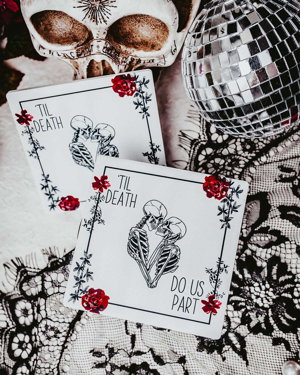 Two square coasters featuring a gothic &#39;Til Death Do Us Part&#39; design with skeletons and red roses, placed on black lace fabric.