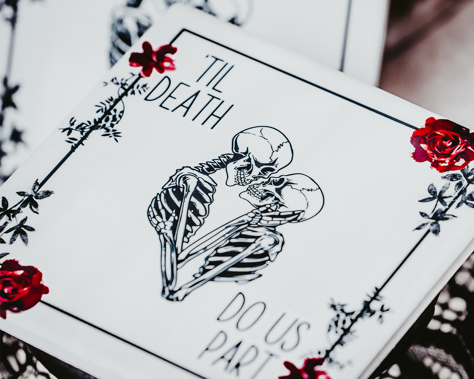 Two square coasters featuring a gothic 'Til Death Do Us Part' design with skeletons and red roses, placed on black lace fabric.