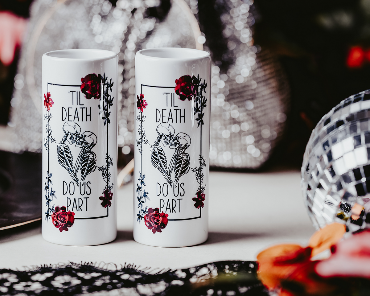 Two white ceramic shot glasses featuring a &#39;Til Death Do Us Part&#39; skeleton design with red roses, perfect for gothic weddings or bridal gifts. Shot glasses are 3oz and showcase a bold, edgy look.