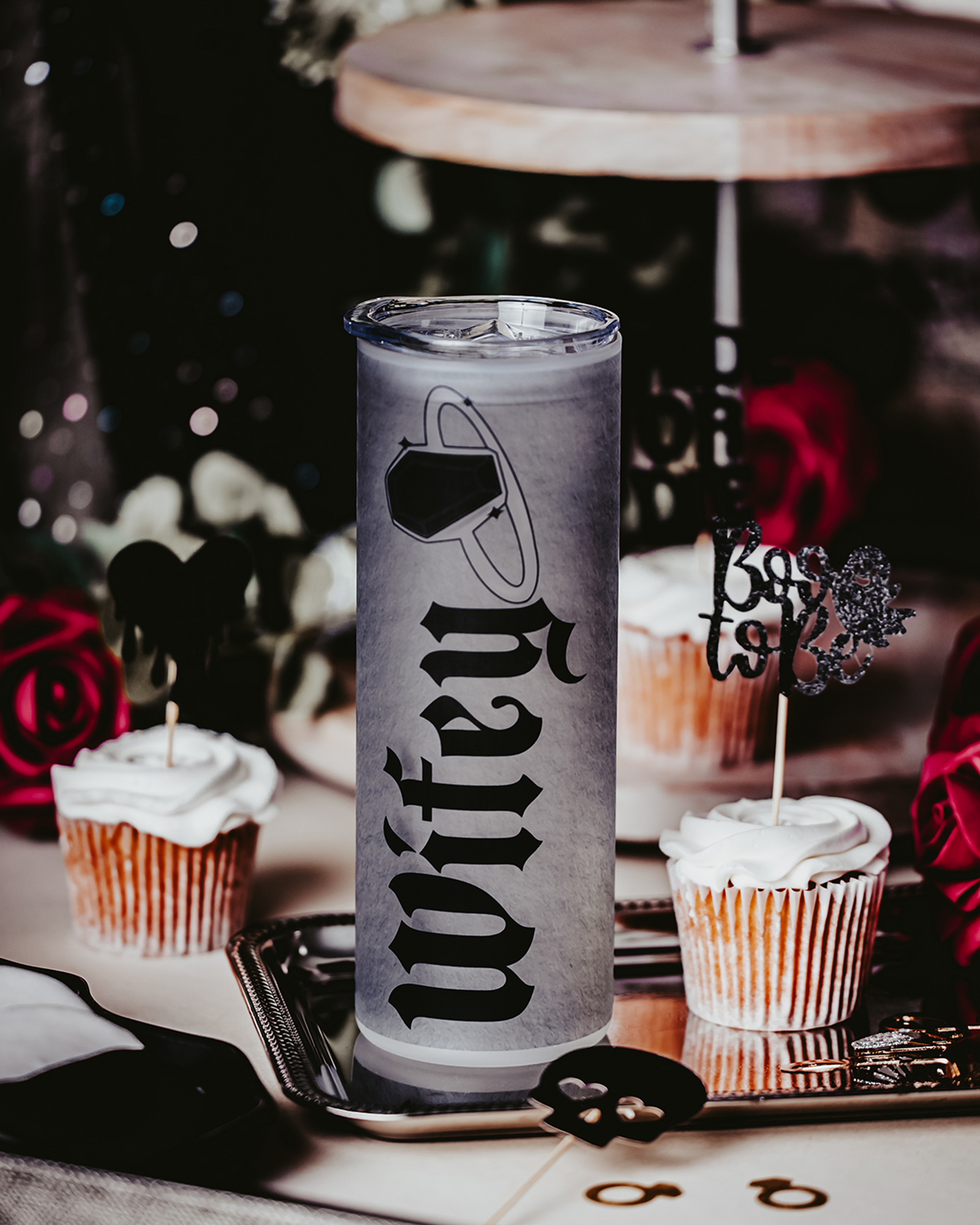 Frosted glass tumbler with gothic black lettering spelling 'Wifey,' accompanied by a coffin ring illustration. The tumbler is lying on its side amidst red roses, cupcakes, and decorative elements, creating a dark, romantic bridal vibe.