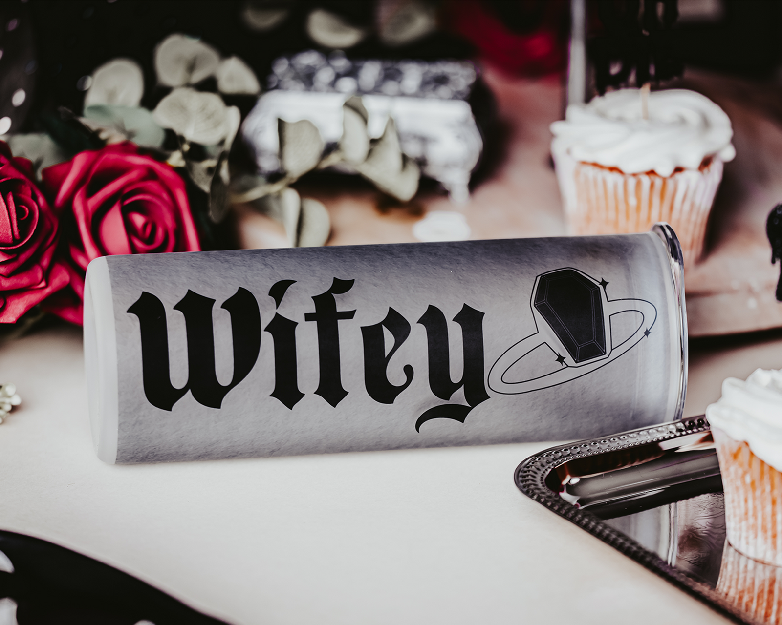 Frosted glass tumbler with gothic black lettering spelling 'Wifey,' accompanied by a coffin ring illustration. The tumbler is lying on its side amidst red roses, cupcakes, and decorative elements, creating a dark, romantic bridal vibe.