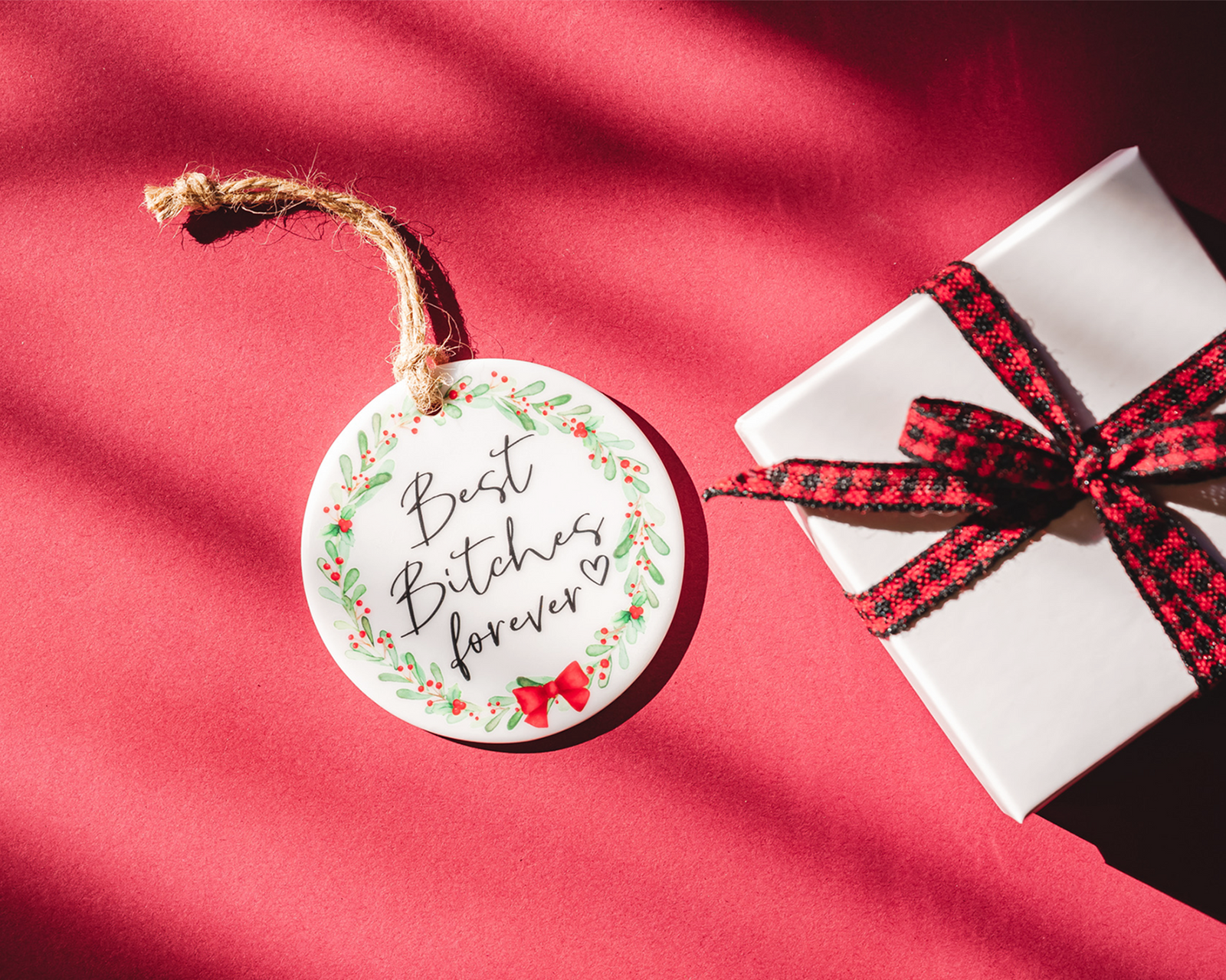 "Round ceramic ornament with the phrase 'Best Bitches Forever' in bold, black script surrounded by a festive holiday wreath of green leaves, red berries, and a red bow, hung with twine against a background of evergreen branches."